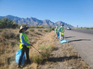 Clear blue skies match the blue ADOT trassh bags cheerfully being filled by volunteers wearing bright yellow safety vests along Oracle Rd. The Catalina mountains ridge line is in the background.